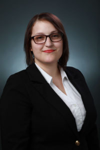 Natasha Crespo is wearing a black suit with a white shirt and glasses. She is smiling in front of a dark blue background