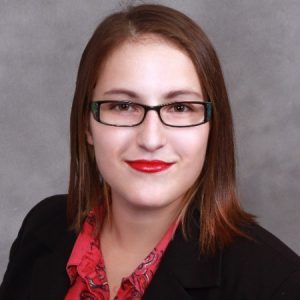 Photo of Natasha Crespo wearing a black suit jacket, a red button-up shirt, and black-framed glasses with red lipstick